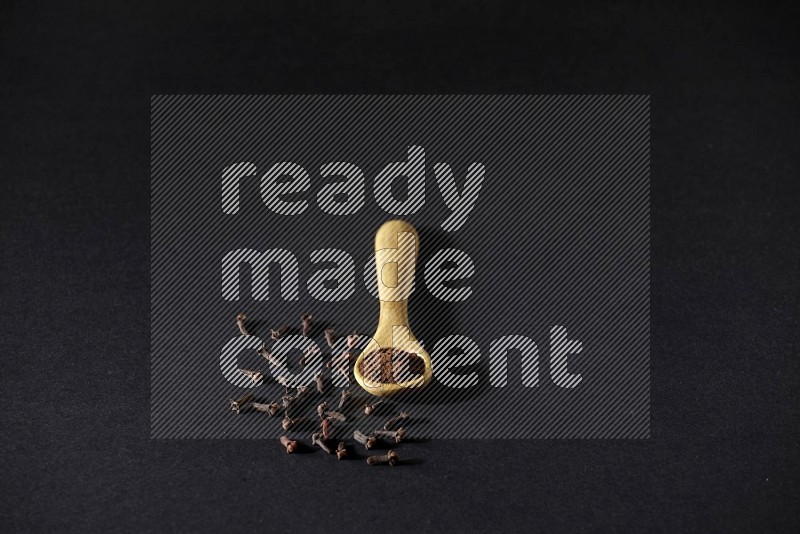 A wooden spoon full of cloves powder with spreaded whole cloves on a black flooring