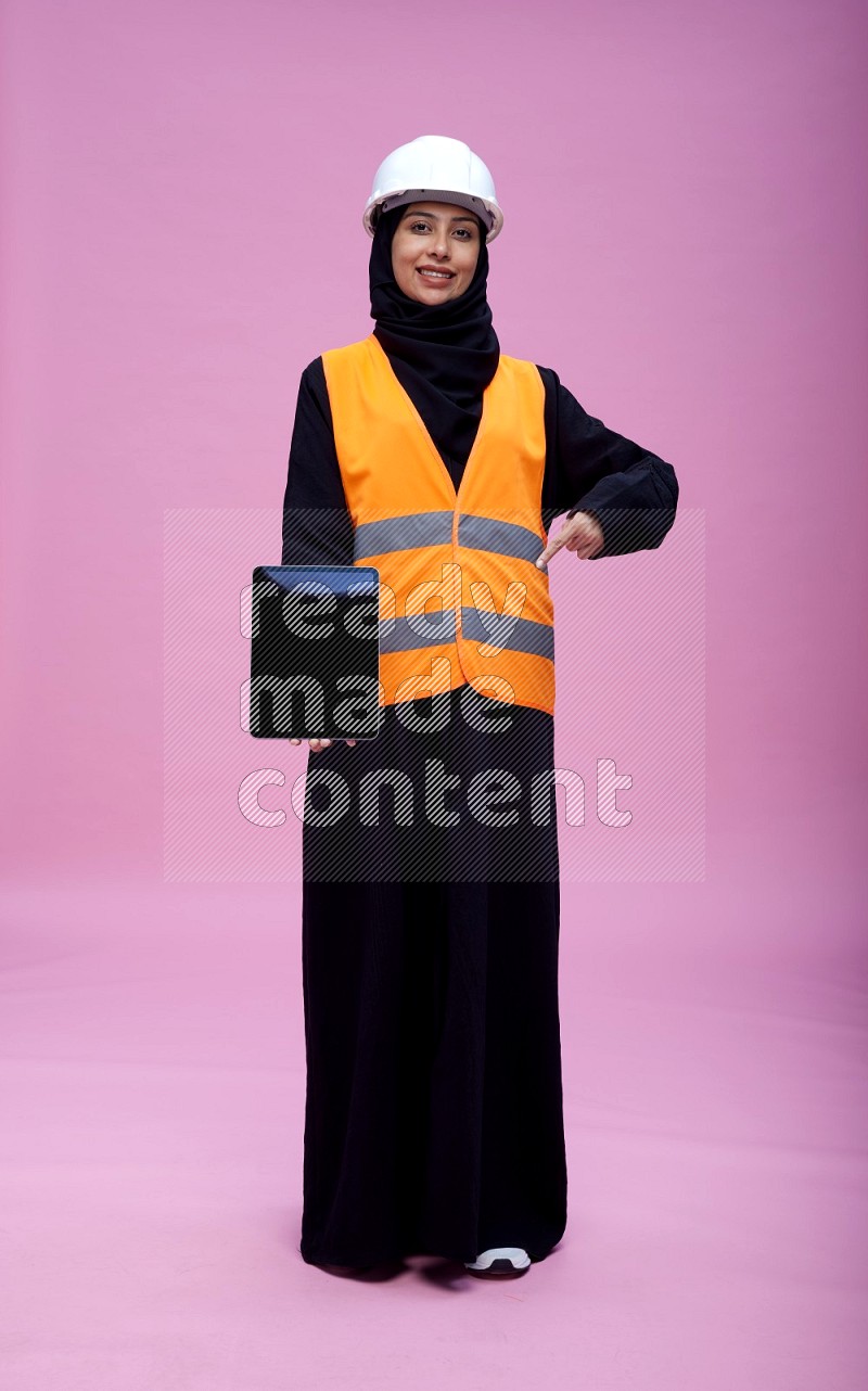 Saudi woman wearing Abaya with engineer vest and helmet standing showing tablet to camera on pink background