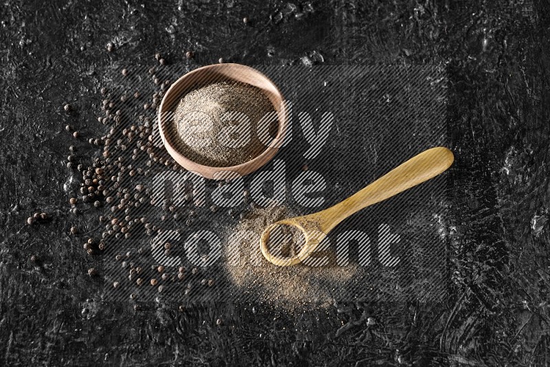 A wooden bowl and a wooden spoon full of black pepper powder with the beads on a textured black flooring