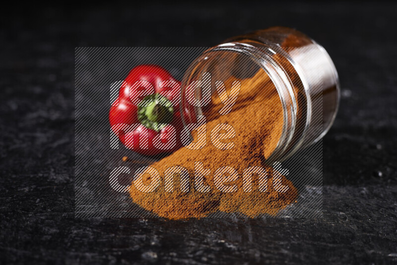 A glass jar full of ground paprika powder flipped with some spilling powder on black background