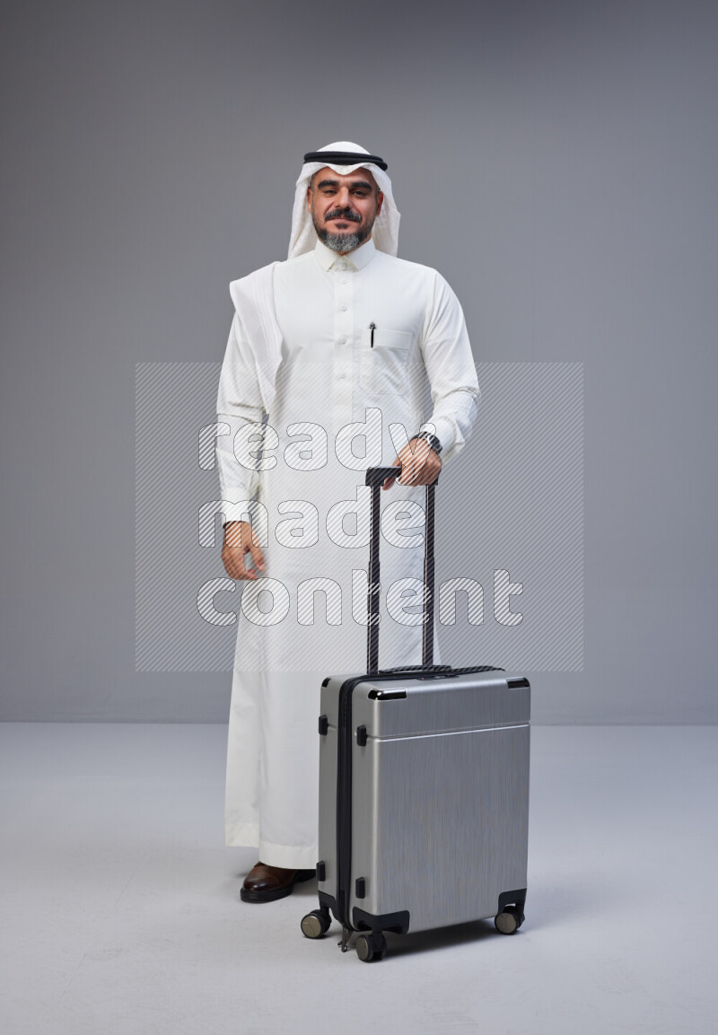 Saudi man wearing Thob and white Shomag standing holding Travel bag on Gray background
