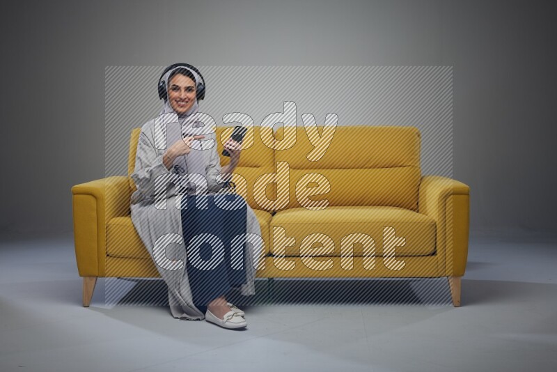 A Saudi woman wearing a light gray Abaya and head scarf sitting on a yellow sofa and using her phone while wearing headphones eye level on a grey background