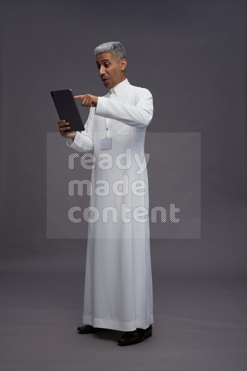 Saudi man wearing thob with neck strap employee badge standing working on tablet on gray background