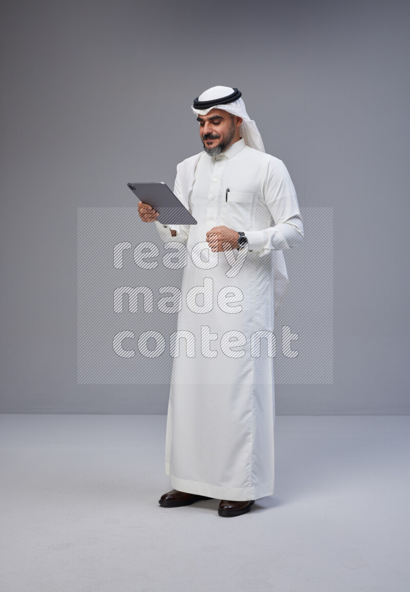 Saudi man Wearing Thob and white Shomag standing working on tablet on Gray background