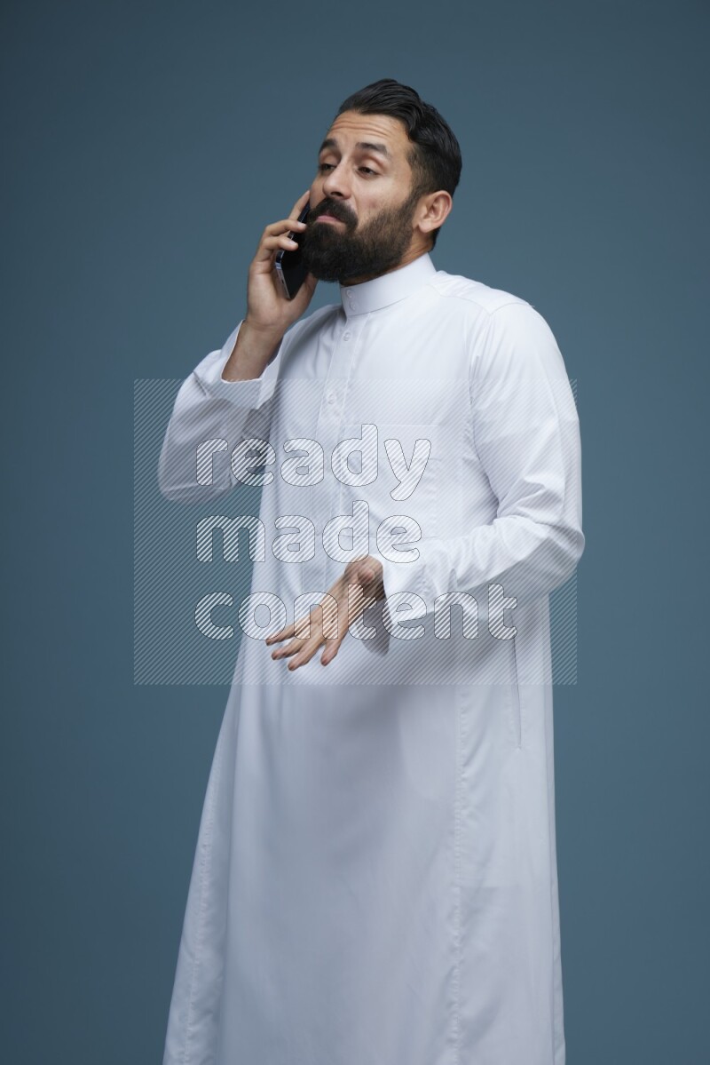 A man having a call on a blue background wearing Saudi Thob