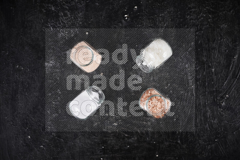 4 glass jars filled with table salt, coarse sea salt, fine himalayan salt and coarse himalayan salt on black background