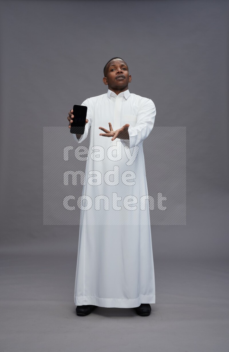 Saudi man wearing thob standing showing phone to camera on gray background