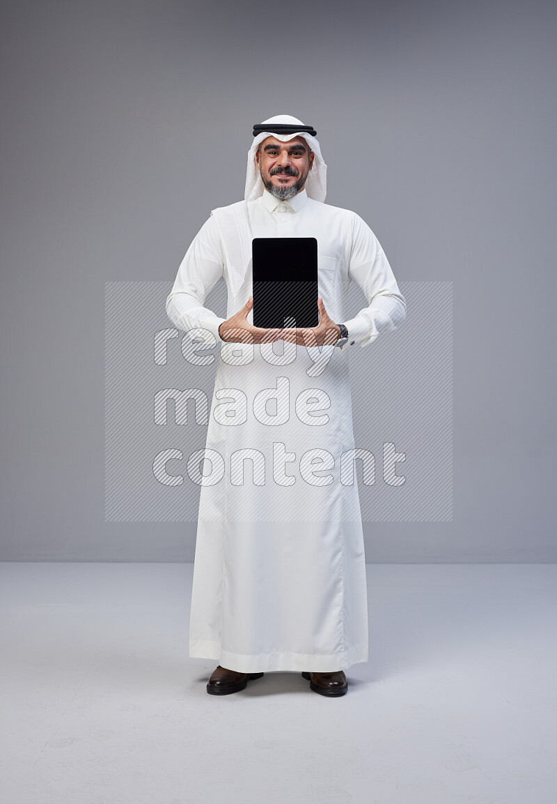 Saudi man Wearing Thob and white Shomag standing showing tablet to camera on Gray background