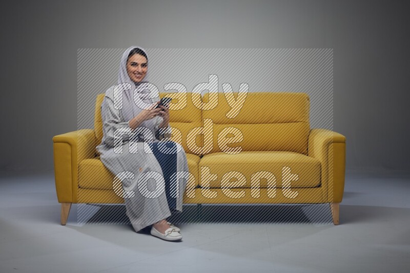 A Saudi woman wearing a light gray Abaya and white head scarf sitting on a yellow sofa and using her phone eye level on a grey background