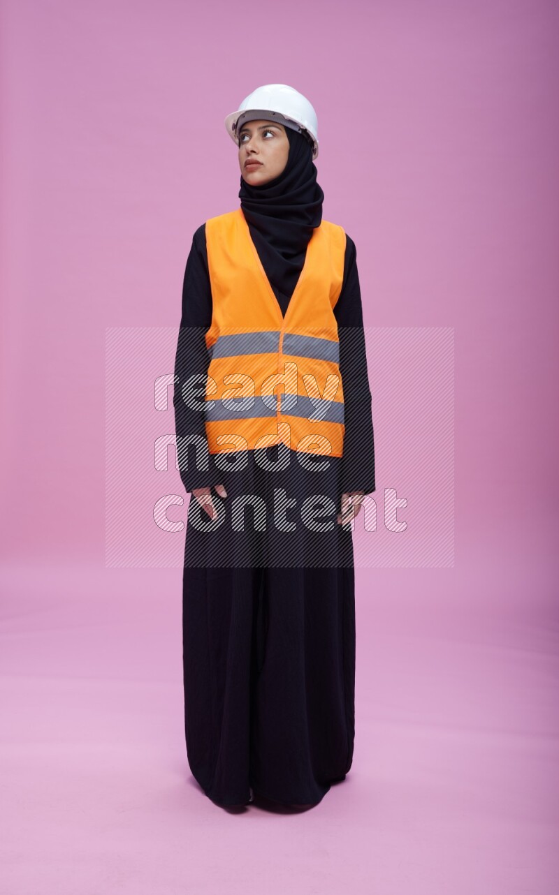 Saudi woman wearing Abaya with engineer vest and helmet standing interacting with the camera on pink background