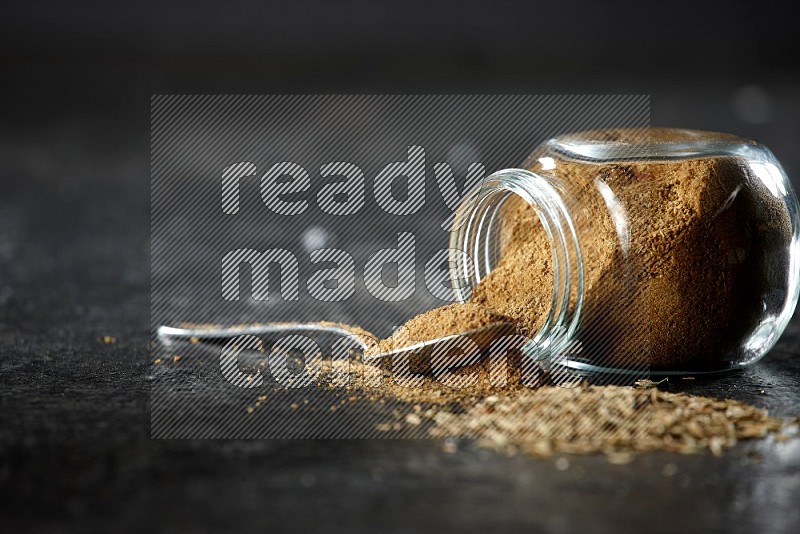 A flipped glass spice jar and a metal spoon full of cumin powder and powder spilled out with cumin seeds on a textured black flooring