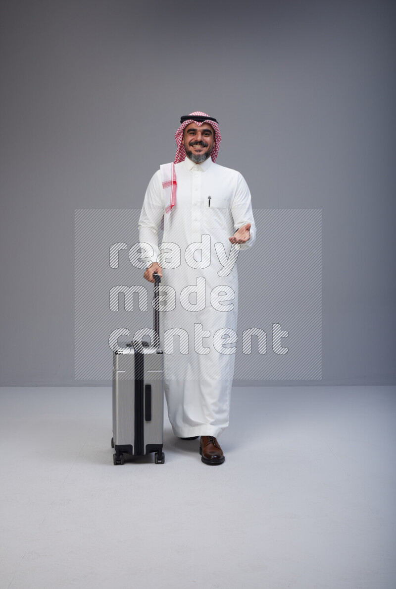 Saudi man wearing Thob and red Shomag standing holding Travel bag on Gray background