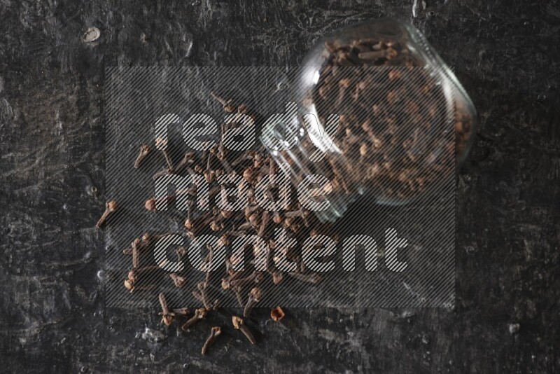 A flipped glass spice jar full of cloves and cloves came out of it on textured black flooring