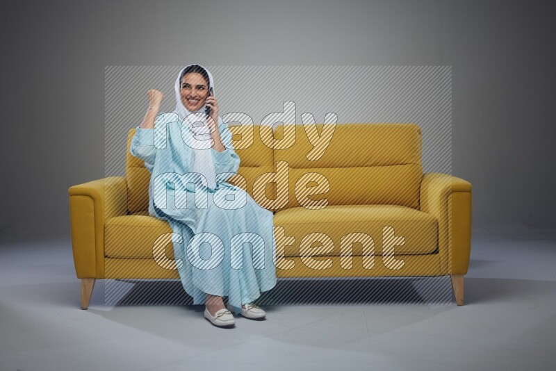 A Saudi woman wearing a light blue Abaya and a white head scarf sitting on a yellow sofa and talking in the phone while making different poses eye level on a grey background