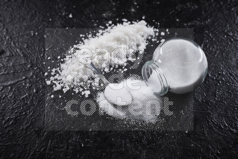 A glass jar full of table salt with some sea salt crystals beside it on a black background