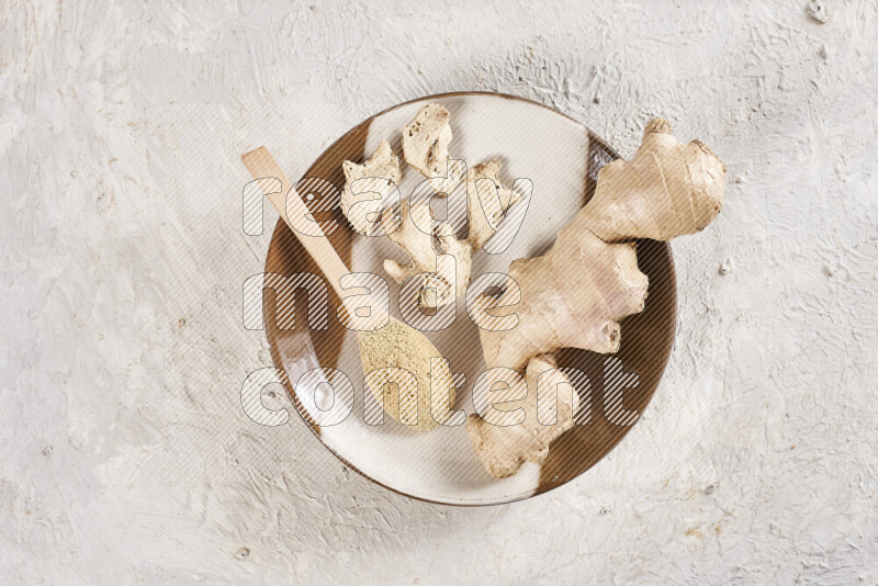 A wooden spoon full of ground ginger powder with fresh and dried ginger, all on a pottery plate on white background