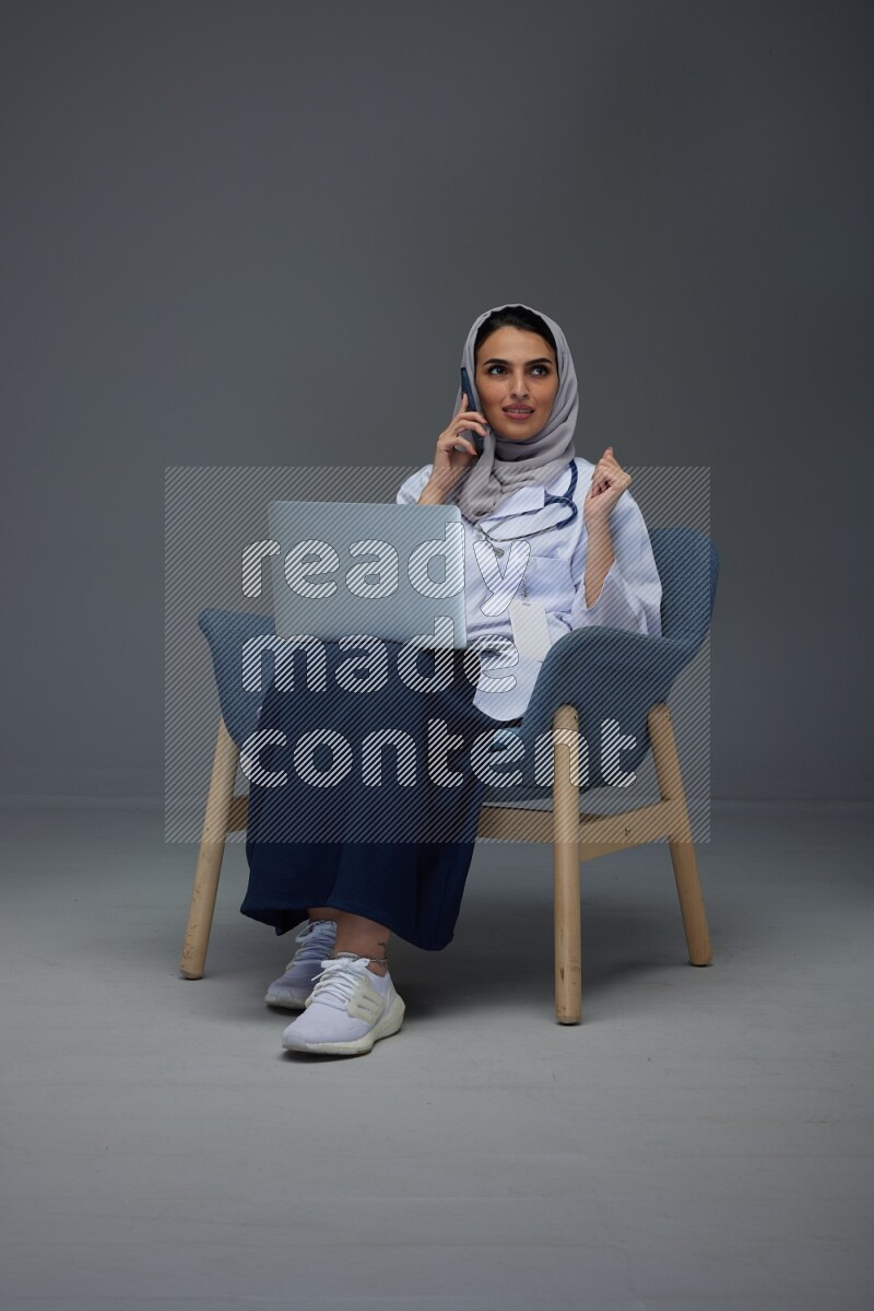 A doctor wearing a light gray head scarf sitting on blue chair on grey background