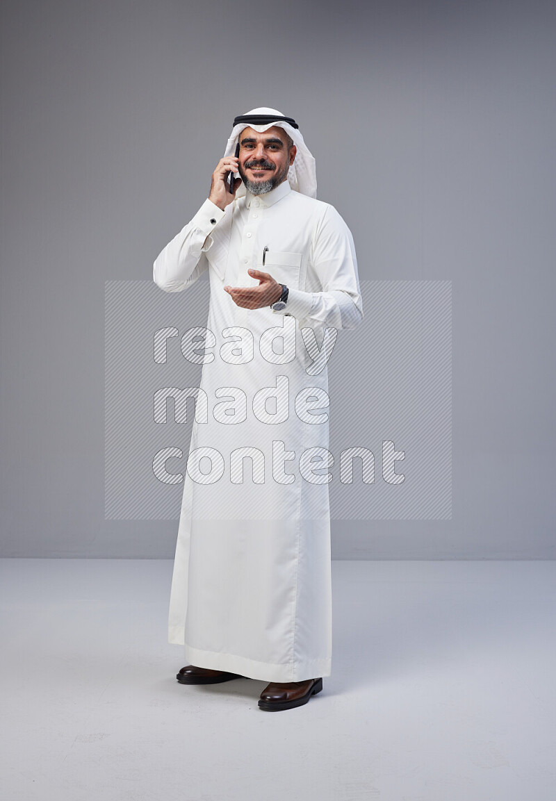 Saudi man Wearing Thob and white Shomag standing talking on phone on Gray background