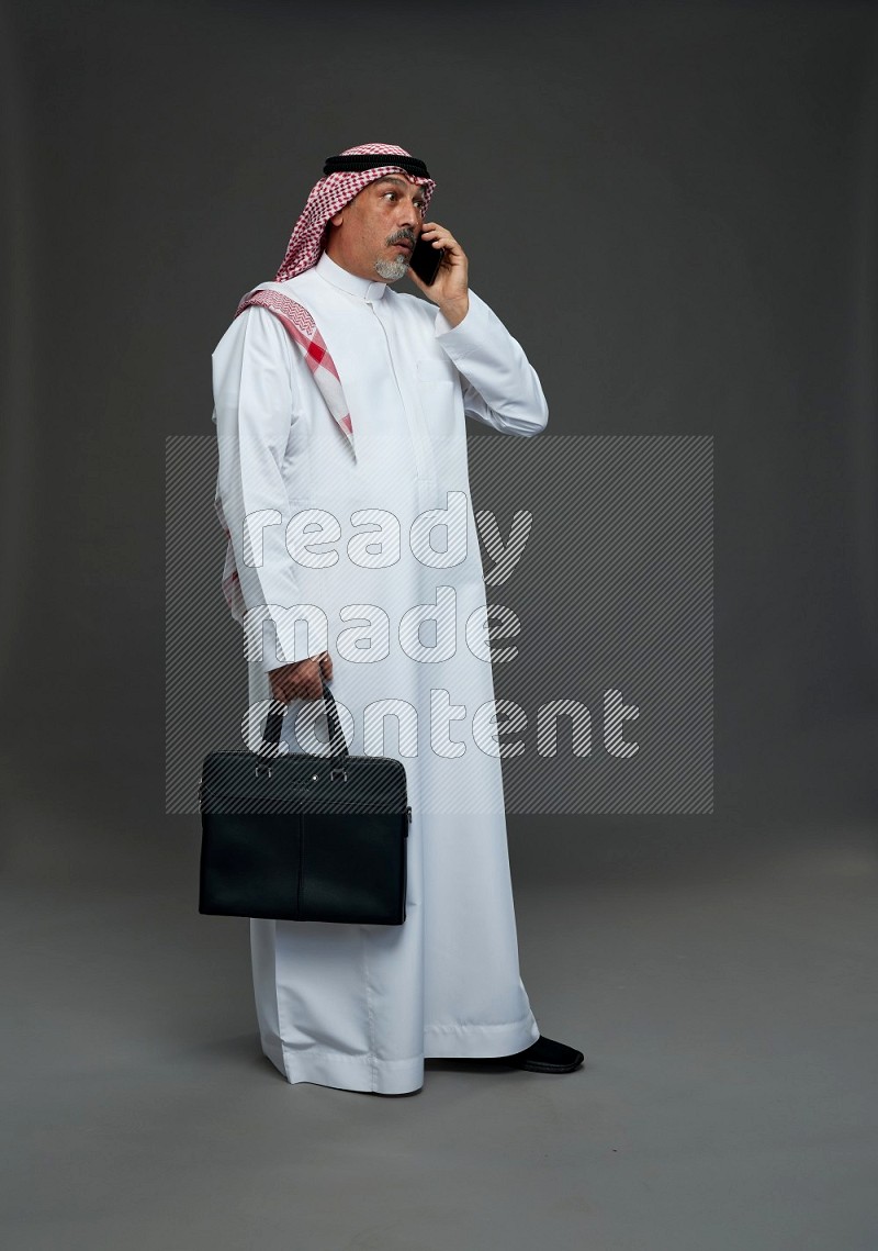 Saudi man with shomag Standing holding bag talking on phone on gray background