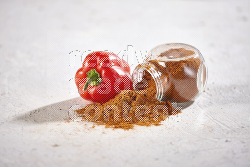 A glass jar full of ground paprika powder flipped with some spilling powder on white background