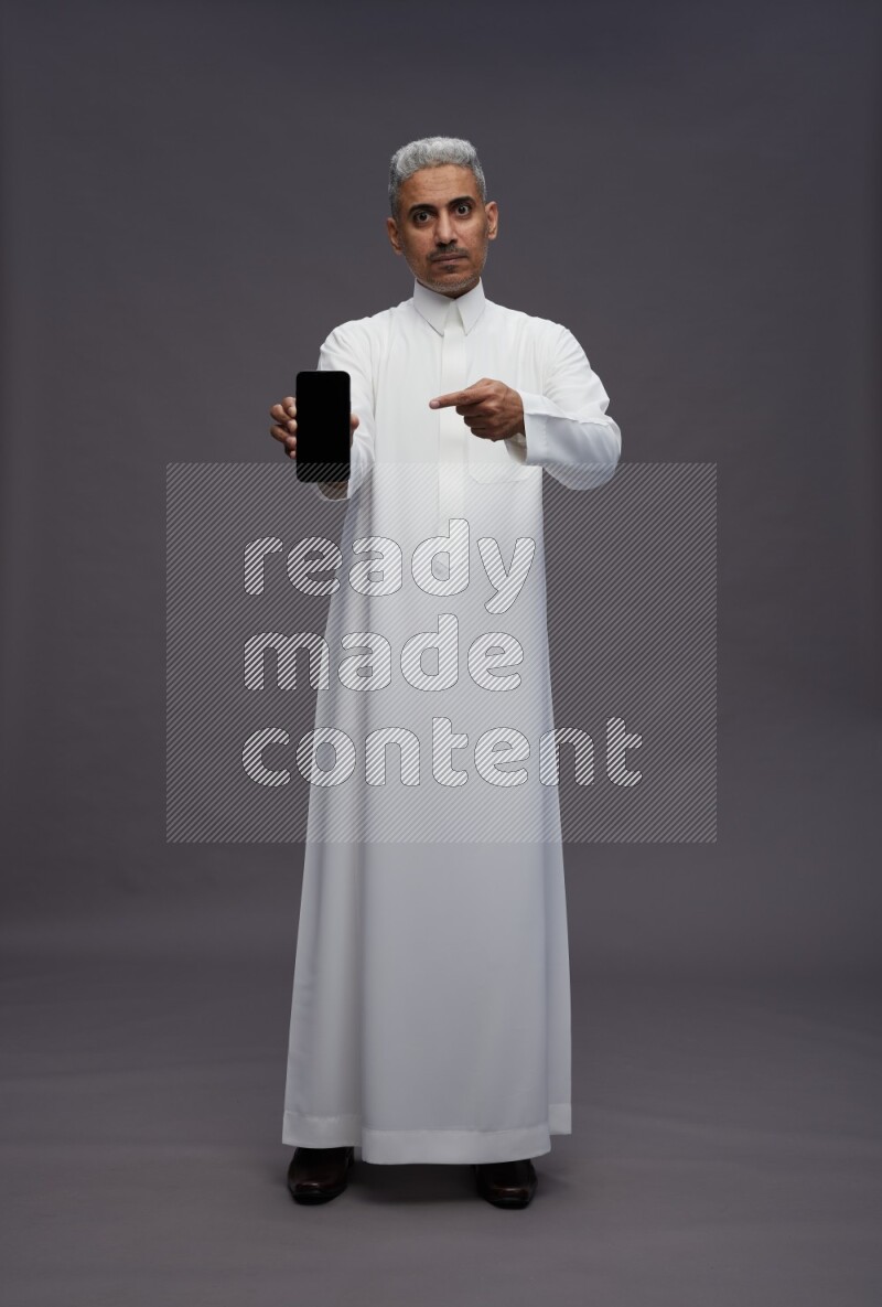 Saudi man wearing thob standing showing phone to camera on gray background