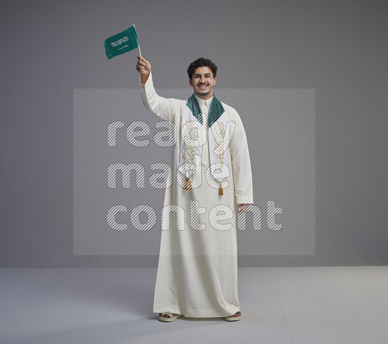 A Saudi man standing wearing thob and saudi flag scarf and holding small saudi flag on gray background