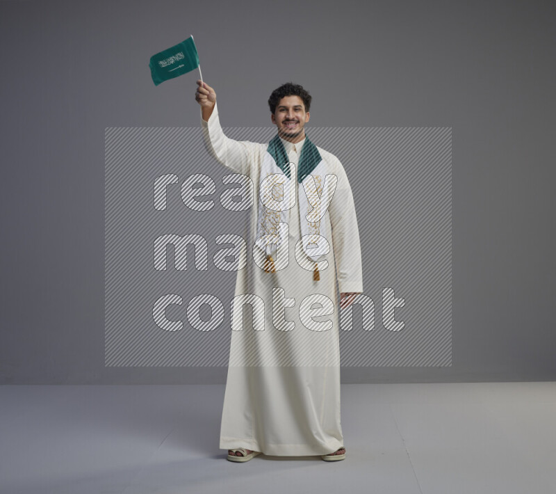 A Saudi man standing wearing thob and saudi flag scarf and holding small saudi flag on gray background
