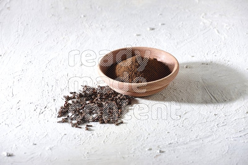A wooden bowl full of cloves powder with whole cloves beside it on a textured white flooring
