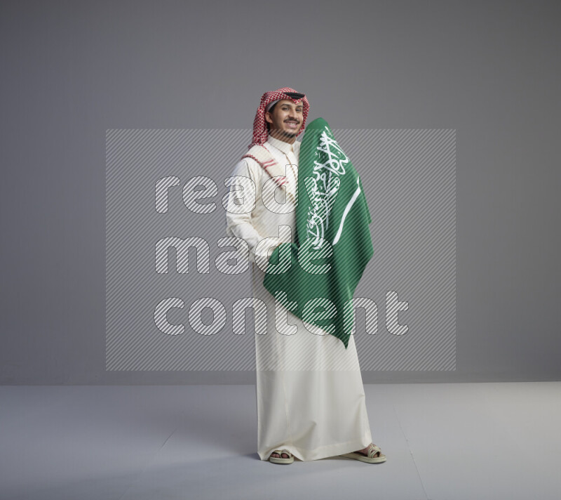 A saudi man standing wearing thob and red shomag holding big saudi flag on gray background