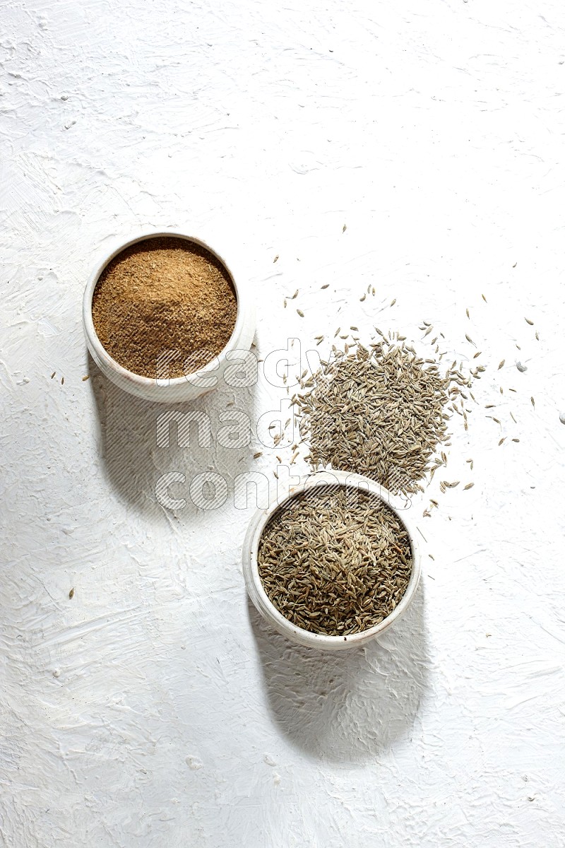 2 beige bowls full of cumin seeds and powder with spilled powder and seeds on textured white flooring