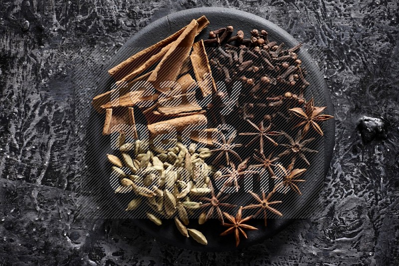 Cinnamon sticks, cloves, star anise and cardamom on a black plate on textured black background