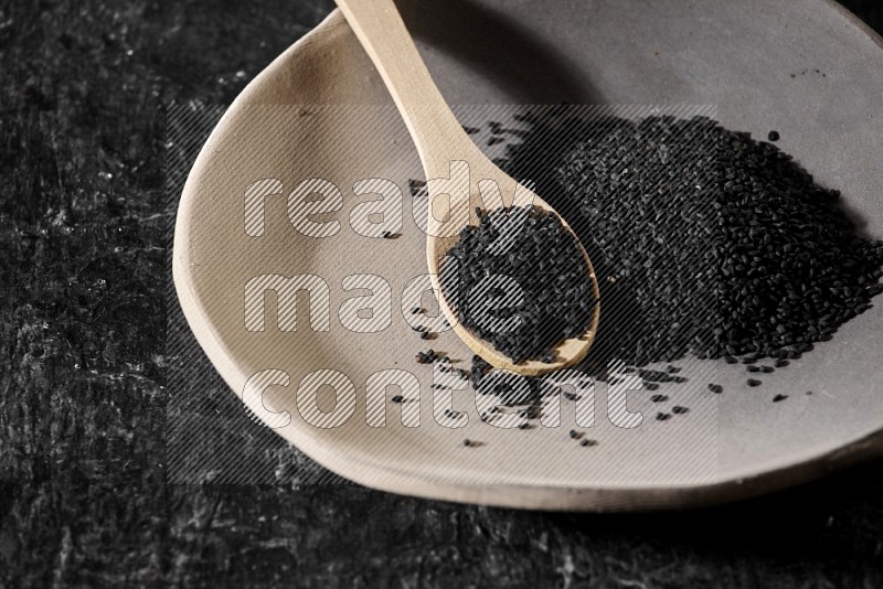 A multicolored pottery plate full of black seeds and wooden spoon full of seeds on a textured black flooring in different angles