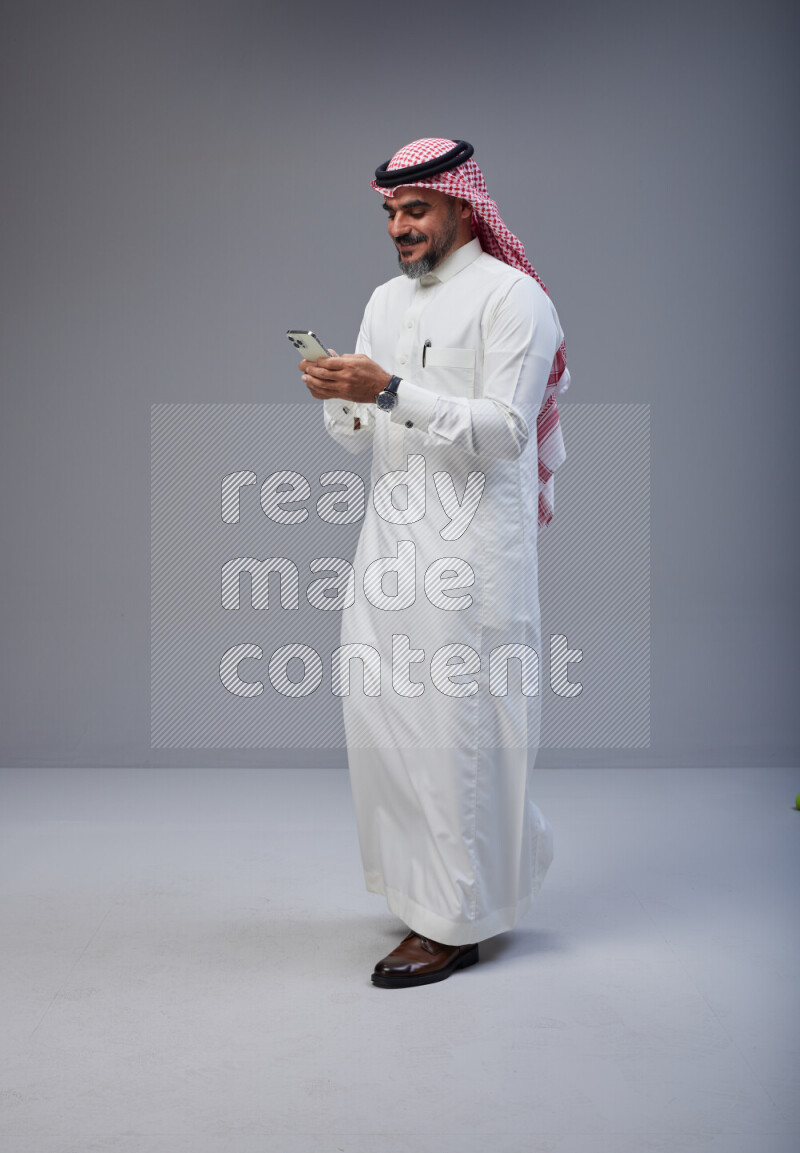 Saudi man Wearing Thob and red Shomag standing texting on phone on Gray background