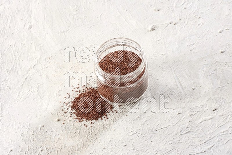 A glass jar full of garden cress seeds with more seeds spread on a textured white flooring
