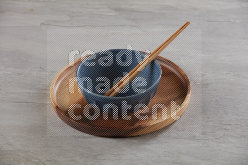Blue pottery round bowl on top of brown wood round plate and wood chopsticks, on grey textured countertop