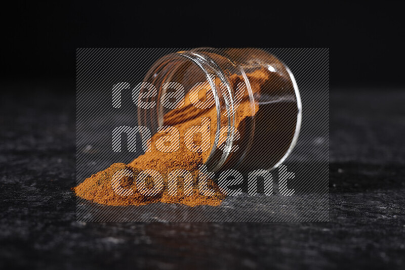 A glass jar full of ground paprika powder flipped with some spilling powder on black background