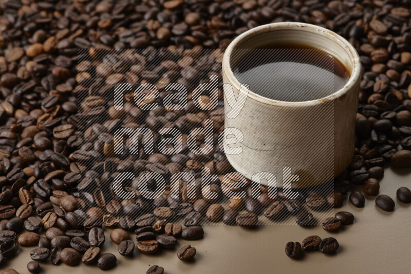 A beige pottery cup of coffee surrounded by roasted coffee beans on beige background