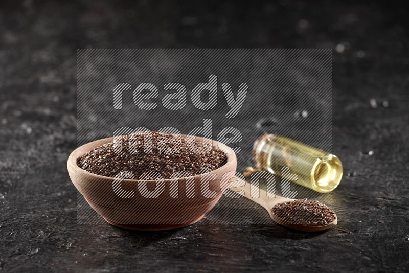 A wooden bowl and spoon full of flaxseeds and a glass bottle of flaxseeds oil on a textured black flooring