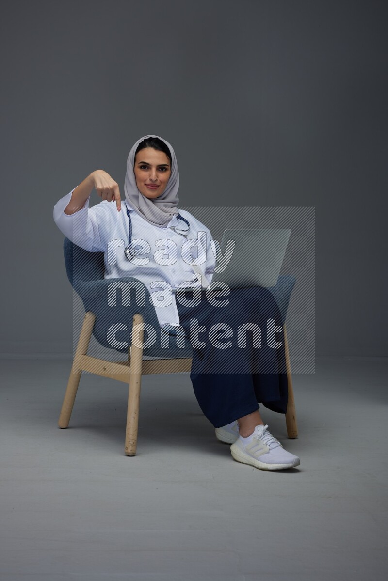 A doctor wearing a light gray head scarf setting on a dark grey chair and using a phone eye level on a grey background