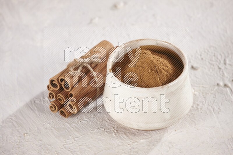 Cinnamon sticks stacked and bounded beside a beige bowl full of cinnamon powder on white background