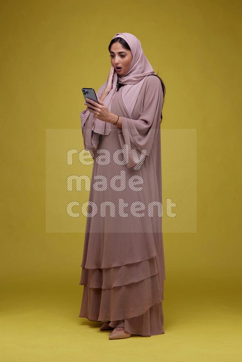 A woman Texting on her phone on a Yellow Background wearing Brown Abaya with Hijab