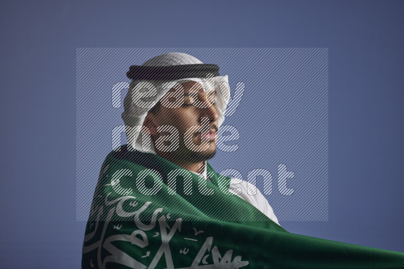 A close-up shot of Saudi man wearing thob and white shomag wrapping big Saudi flag on gray background