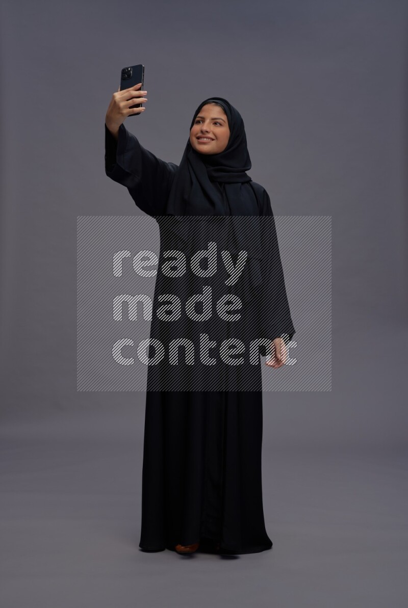 Saudi woman wearing Abaya standing taking selfie on gray background