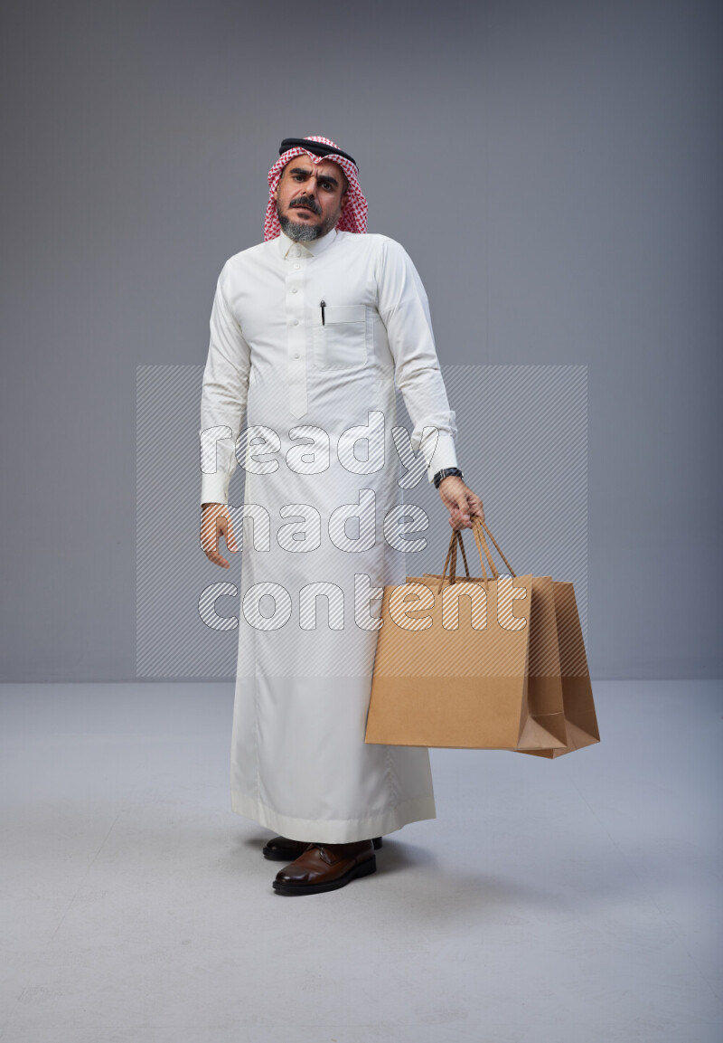 Saudi man Wearing Thob and red Shomag standing holding shopping bag on Gray background