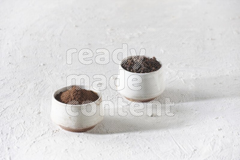2 Beige ceramic bowls, one full of cloves and the other full of powder on textured white flooring