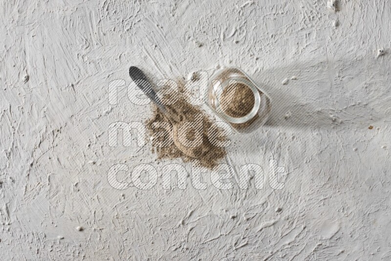 A glass spice jar full of black pepper powder and metal spoon full of it on textured white flooring