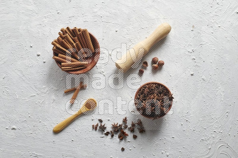 A wooden bowl full of cinnamon sticks and a wooden spoon with cinnamon powder are neatly placed beside a bowl of star anise on a textured white background
