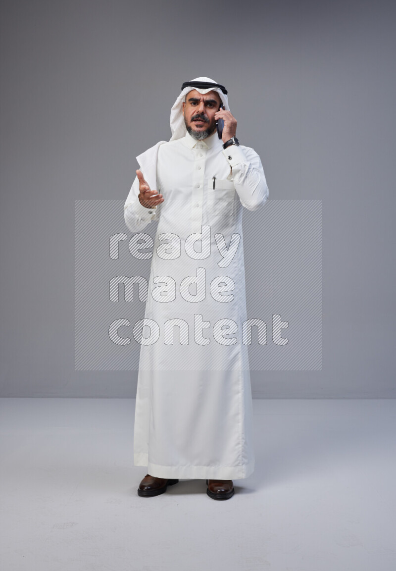 Saudi man Wearing Thob and white Shomag standing talking on phone on Gray background