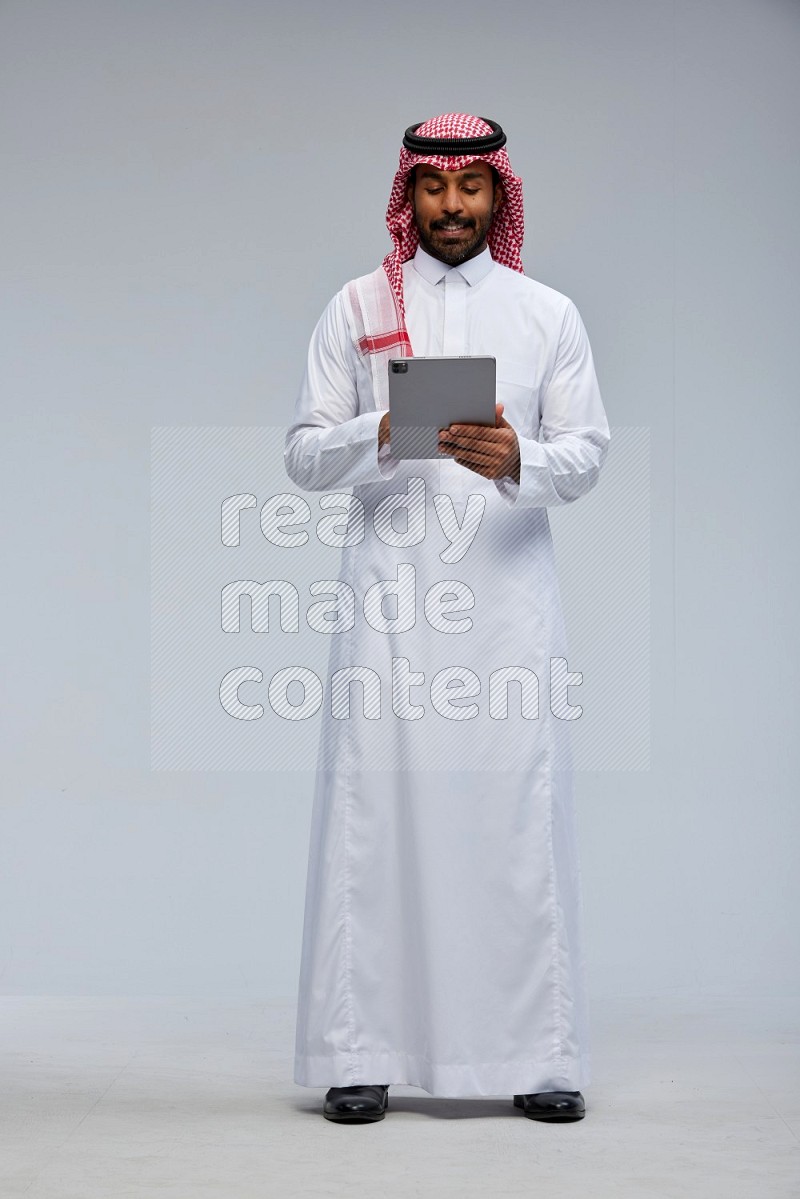 Saudi man Wearing Thob and shomag standing working on tablet on Gray background