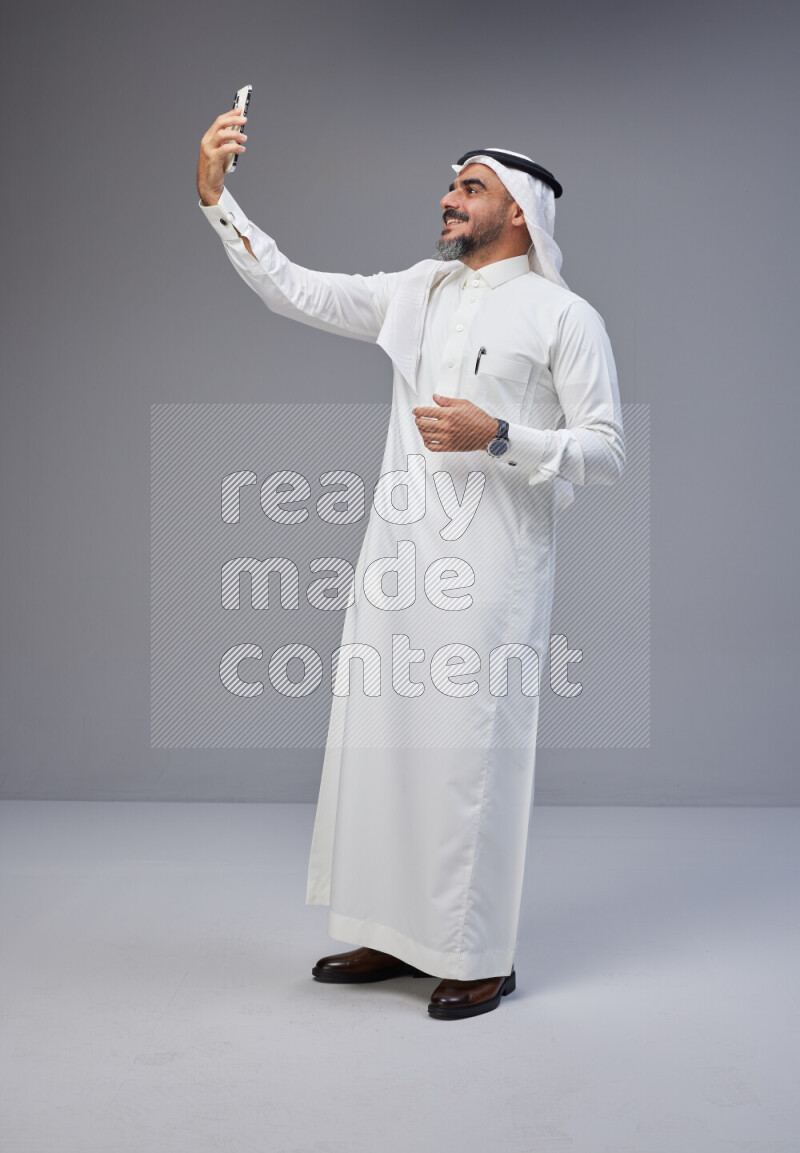Saudi man Wearing Thob and white Shomag standing taking selfie on Gray background